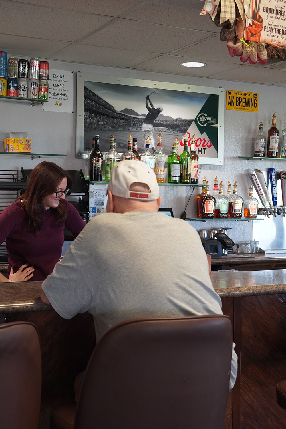 Bartender stands behind a bar counter with liquor bottles on shelves and a patron seated at the bar.